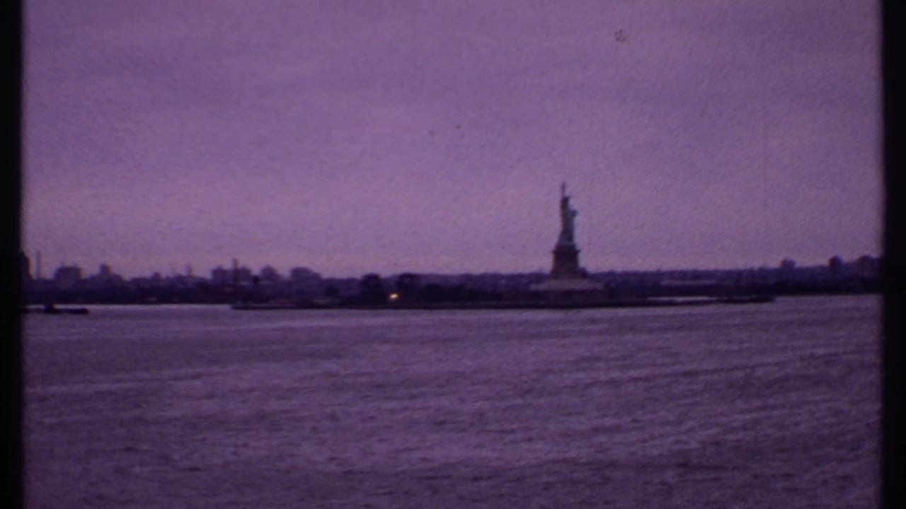 Distant Statue of Liberty across harbor water under a purple sky.