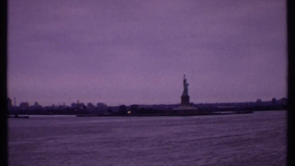 Distant 1969 8mm view of the Statue of Liberty from a boat, framed by water, skyline haze, and film gate edges.