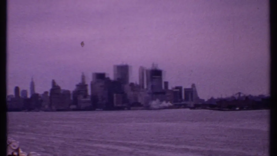 1969 8mm frame of the New York City skyline across harbor water, softened by haze, grain, and purple color shift.