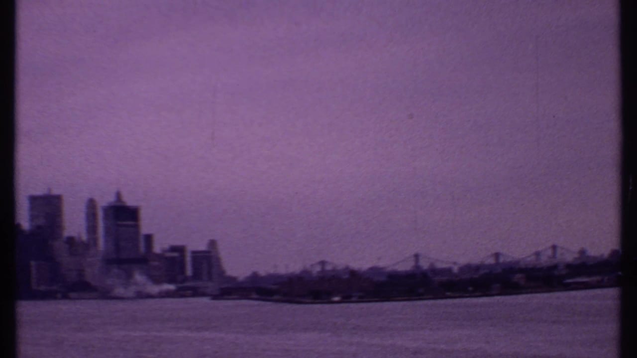 1969 8mm frame showing a distant urban skyline across open water with purple tint and soft film grain.