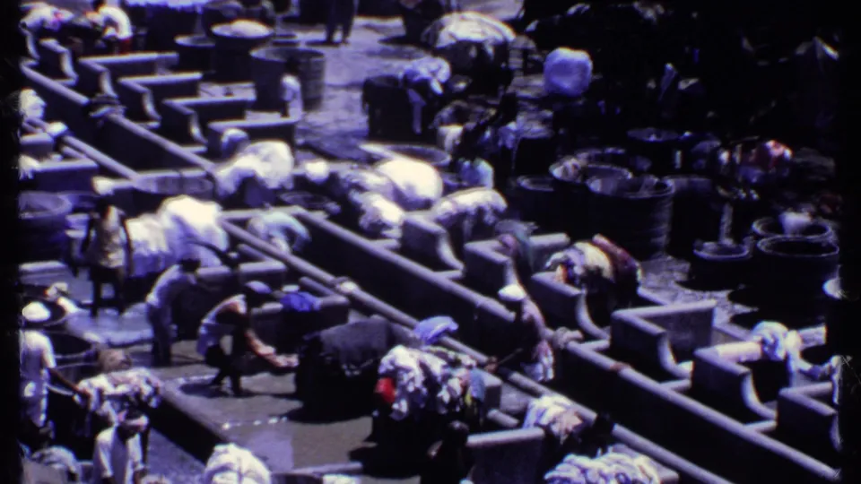 1969 India 8mm frame showing people bending over concrete wash troughs with tubs and laundry bundles around them.