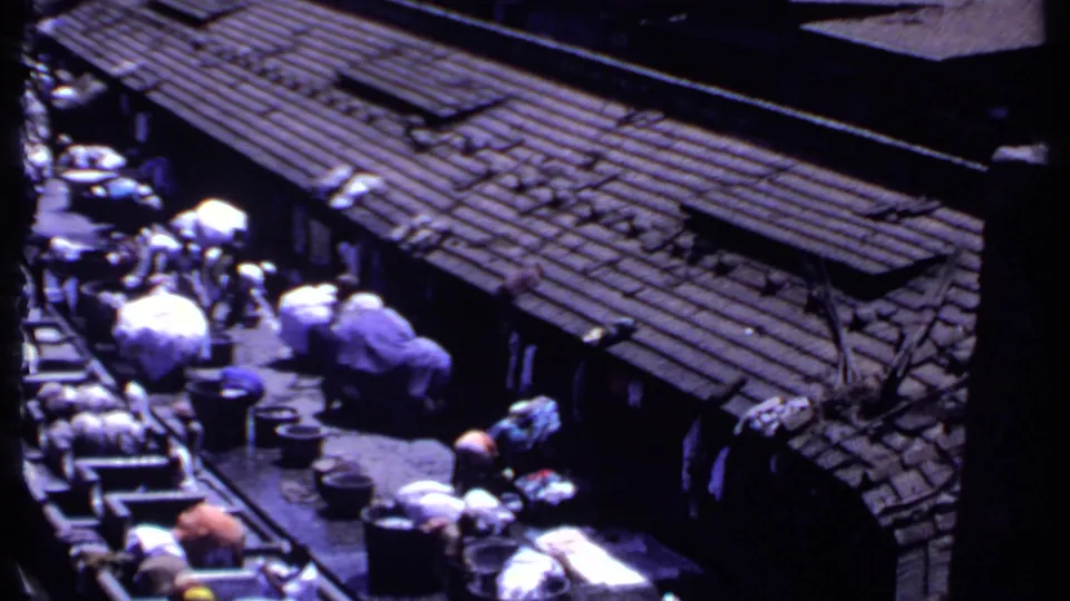 1969 India 8mm frame showing corrugated roofs, people, tubs, and laundry bundles near outdoor wash troughs.