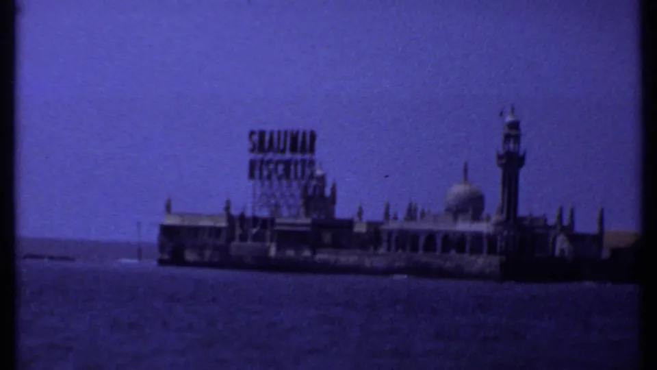 Distant waterfront view in India with a sign-topped building, domed roofline, tower, and open water in the foreground.