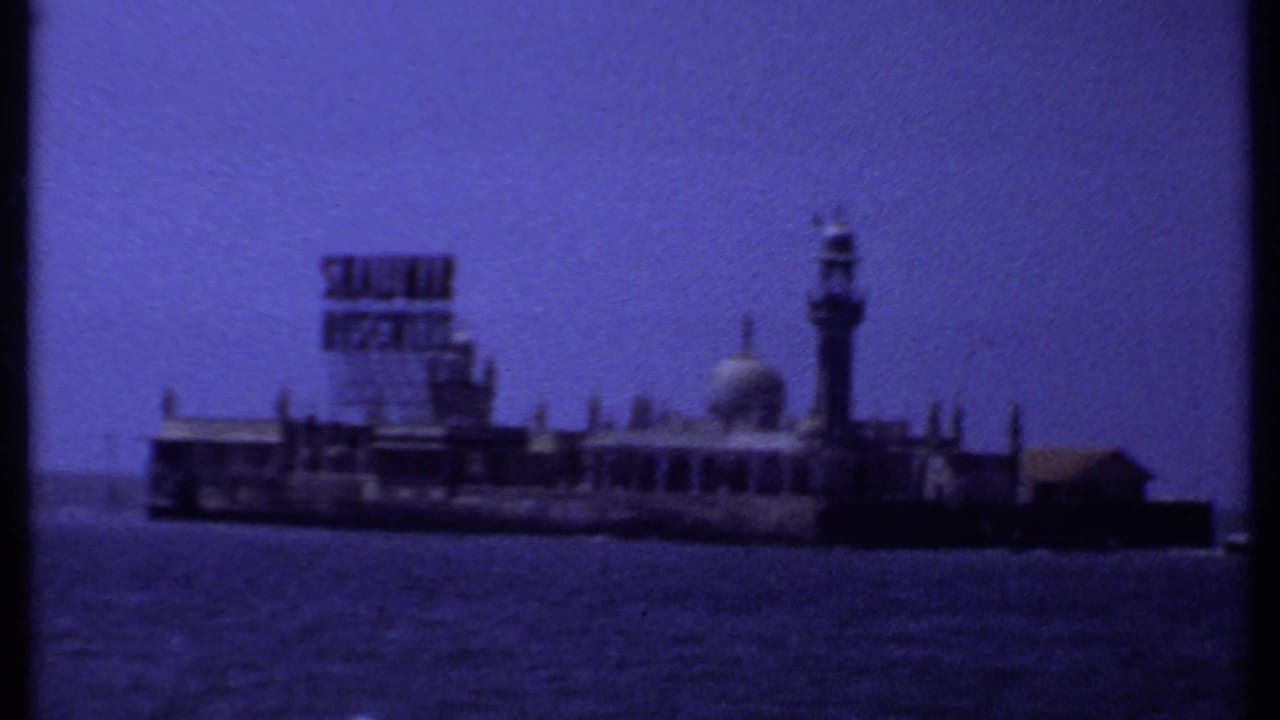 Small offshore building complex with billboard, dome, minaret, sea, and blue sky.