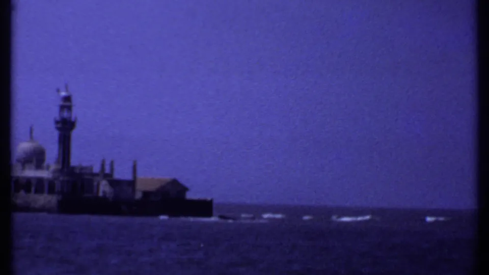 1969 India 8mm frame of the island building complex with a dome, minaret, low structures, sea, and strong blue-purple film cast.