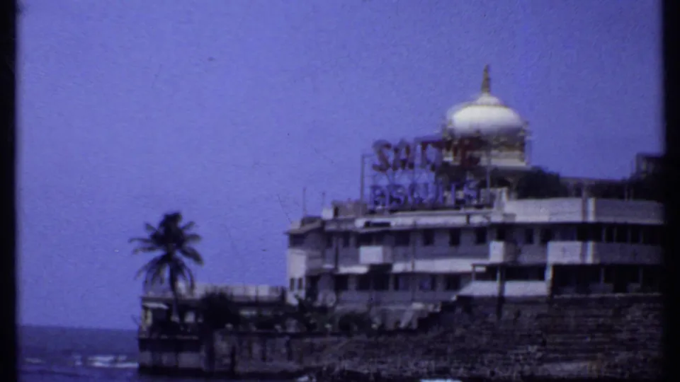 1969 India 8mm frame with the domed Sathe Biscuits building beside the shoreline, with a palm tree and ocean visible.