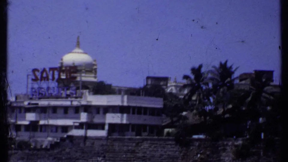 1969 India 8mm frame showing the coastal landmark with rooftop sign, white dome, palm trees and stone shoreline.