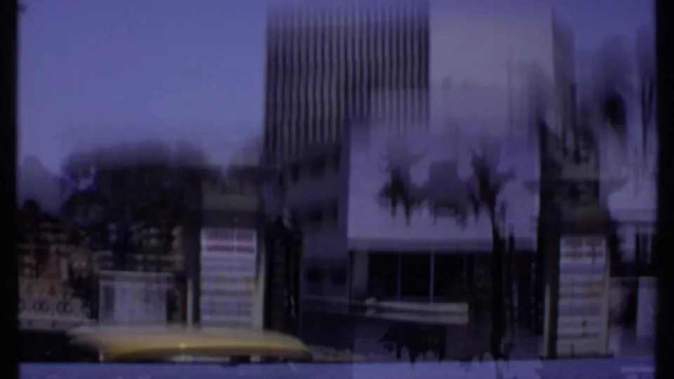 Motion-blurred 1969 India 8mm frame showing the building exterior, palm trunks, entrance area, and the edge of a passing vehicle roof.