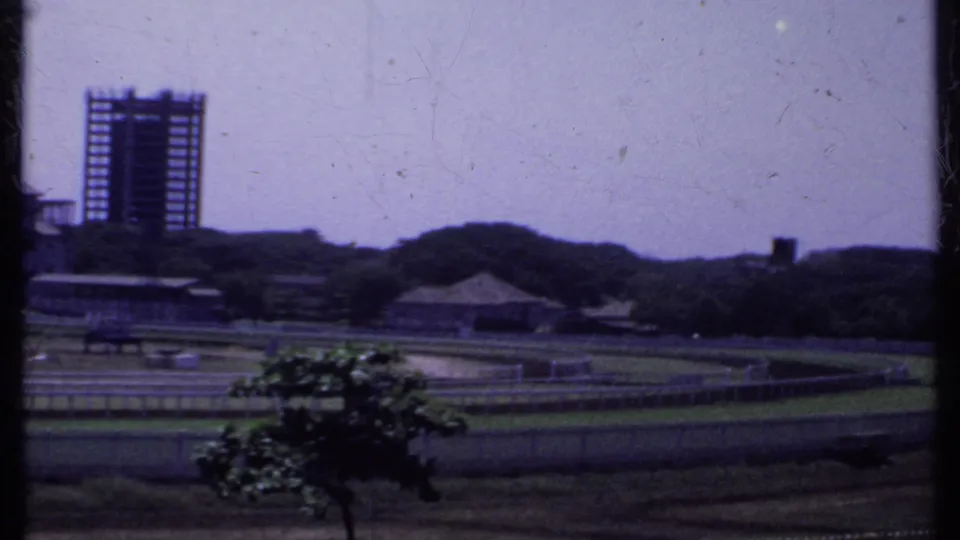 1969 India 8mm frame showing an open oval grounds or racetrack area, distant modern building, trees, low hills, and hazy sky.