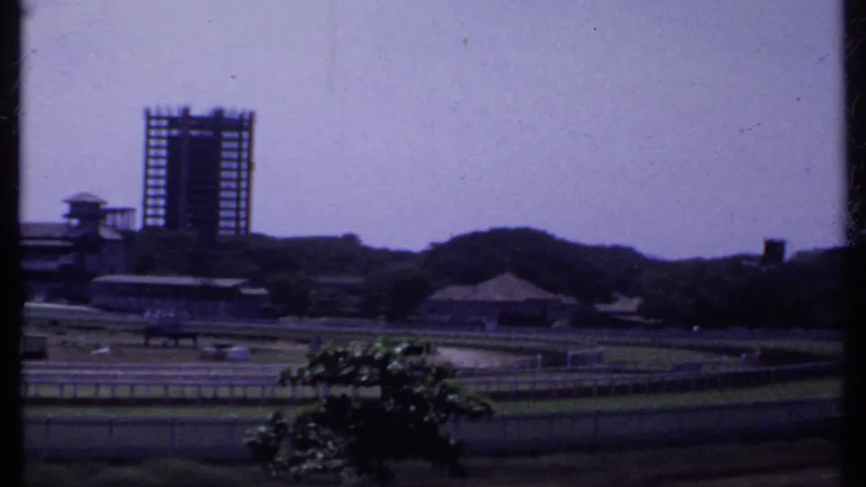 1969 India 8mm frame showing a skeletal high-rise rising behind low buildings, trees, lawns, and rail-lined grounds.
