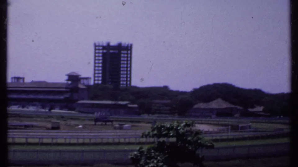 Distant 1969 India view of an unfinished office building frame above low structures, trees, and rail-lined grounds.