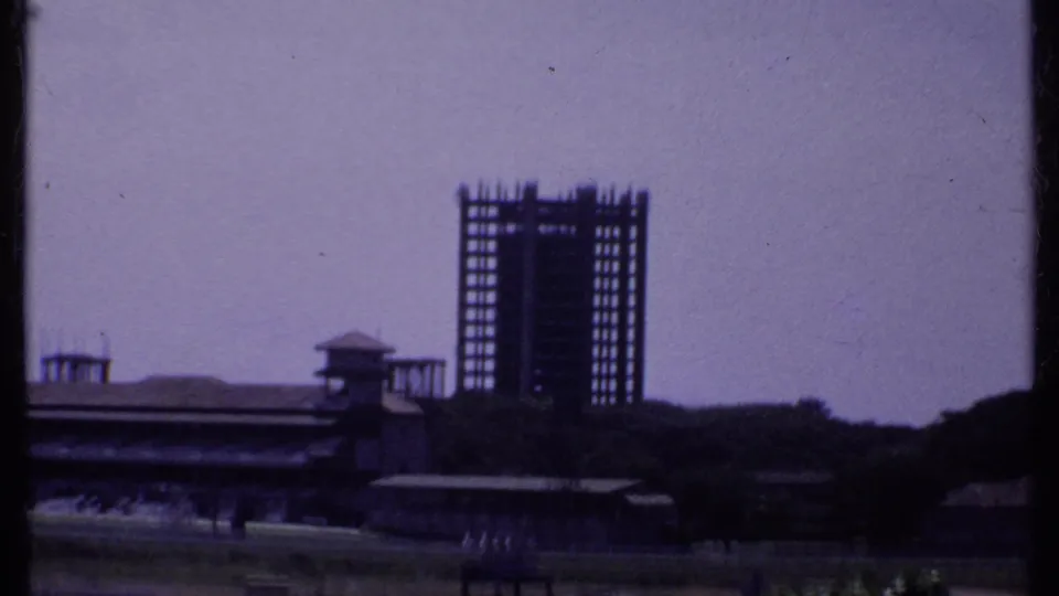 1969 India 8mm still with an unfinished high-rise frame behind a low structure, railings, grass, and a hazy sky.