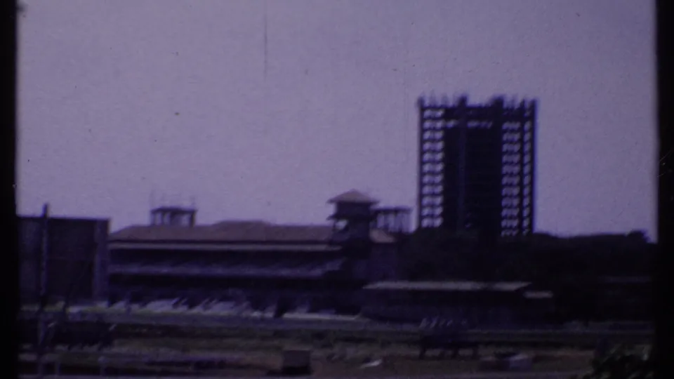 1969 India exterior frame showing the skeletal office building at right, with low buildings, rail-lined grounds, and trees below.