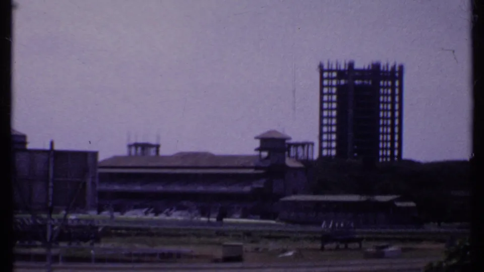 1969 India home-movie frame showing a distant grandstand complex and a taller building frame under construction.