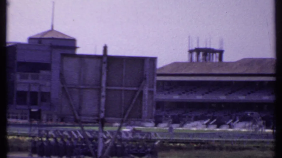 1969 India home-movie view of a boxy stadium-side structure and adjacent covered grandstand across the racetrack grounds.