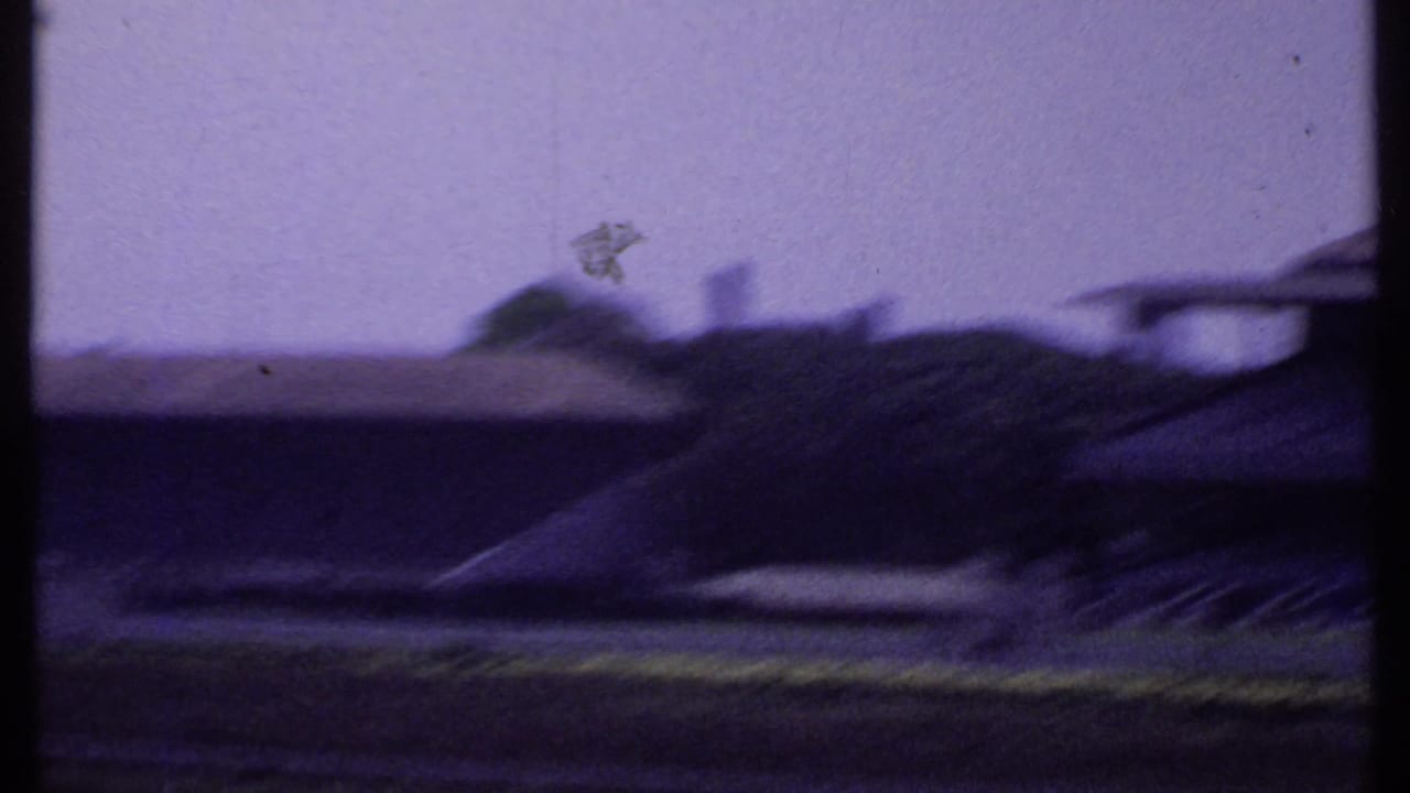 Motion-blurred 8mm frame from the pan across an Indian racetrack, with low buildings, open grounds, and distant rooflines.