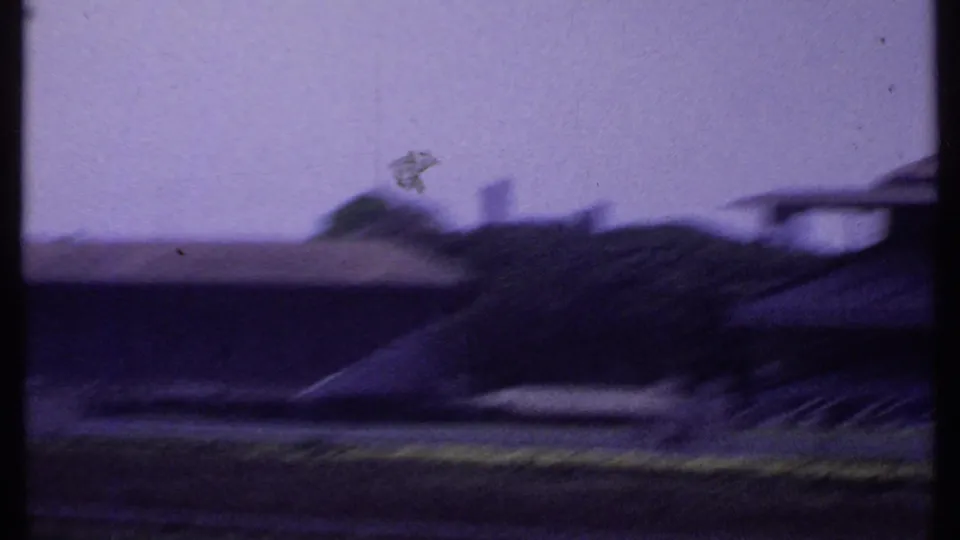 Motion-blurred 8mm frame from the pan across an Indian racetrack, with low buildings, open grounds, and distant rooflines.