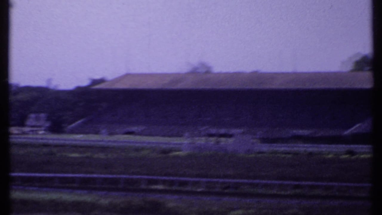 1969 India 8mm frame showing the empty racetrack and a long covered grandstand or stand building under a pale sky.