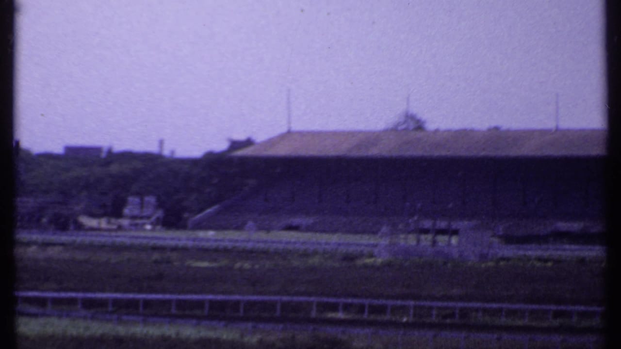 Distant stadium-like building across open ground, seen through a dark vertical frame edge.