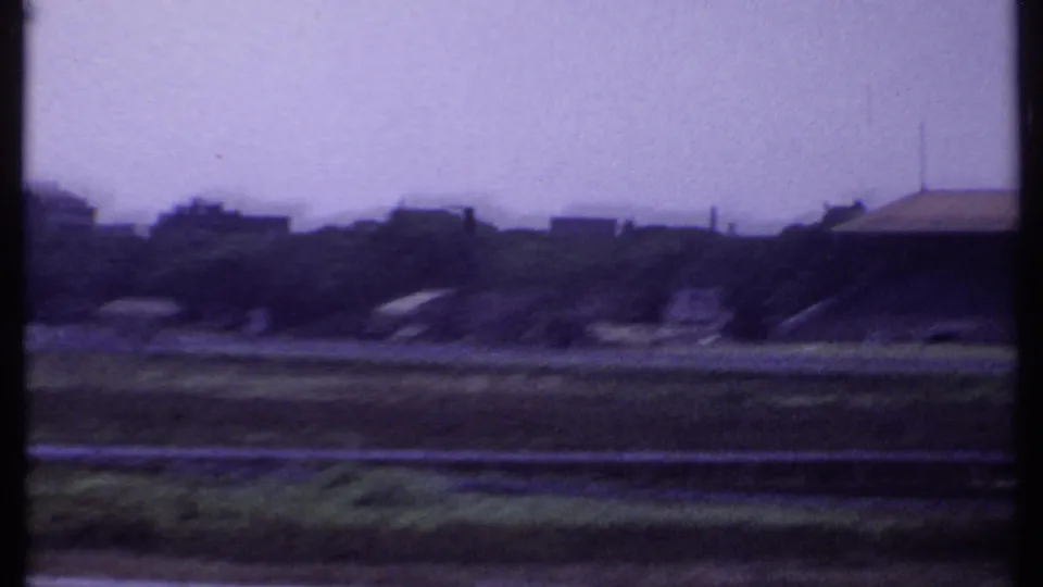 Soft 8mm frame of open grounds in India with distant low buildings, trees, and a vertical dark frame edge.
