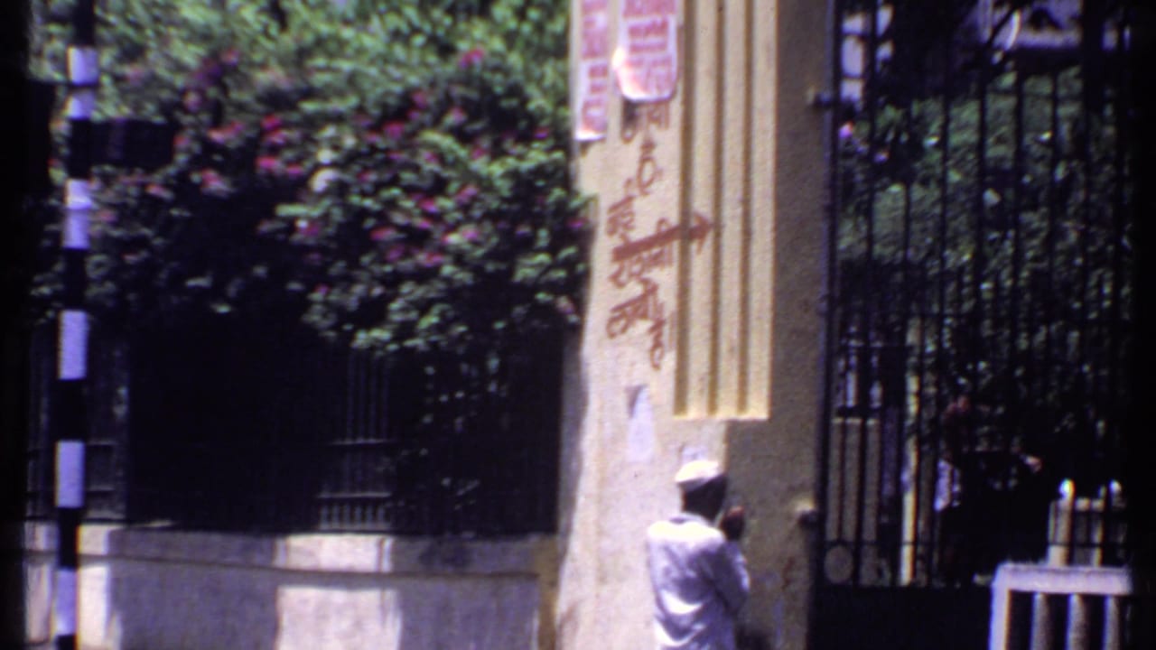 1969 India street-edge view with an iron gate, striped post, dense shrubs, painted pillar, and a person partly visible near the entrance.