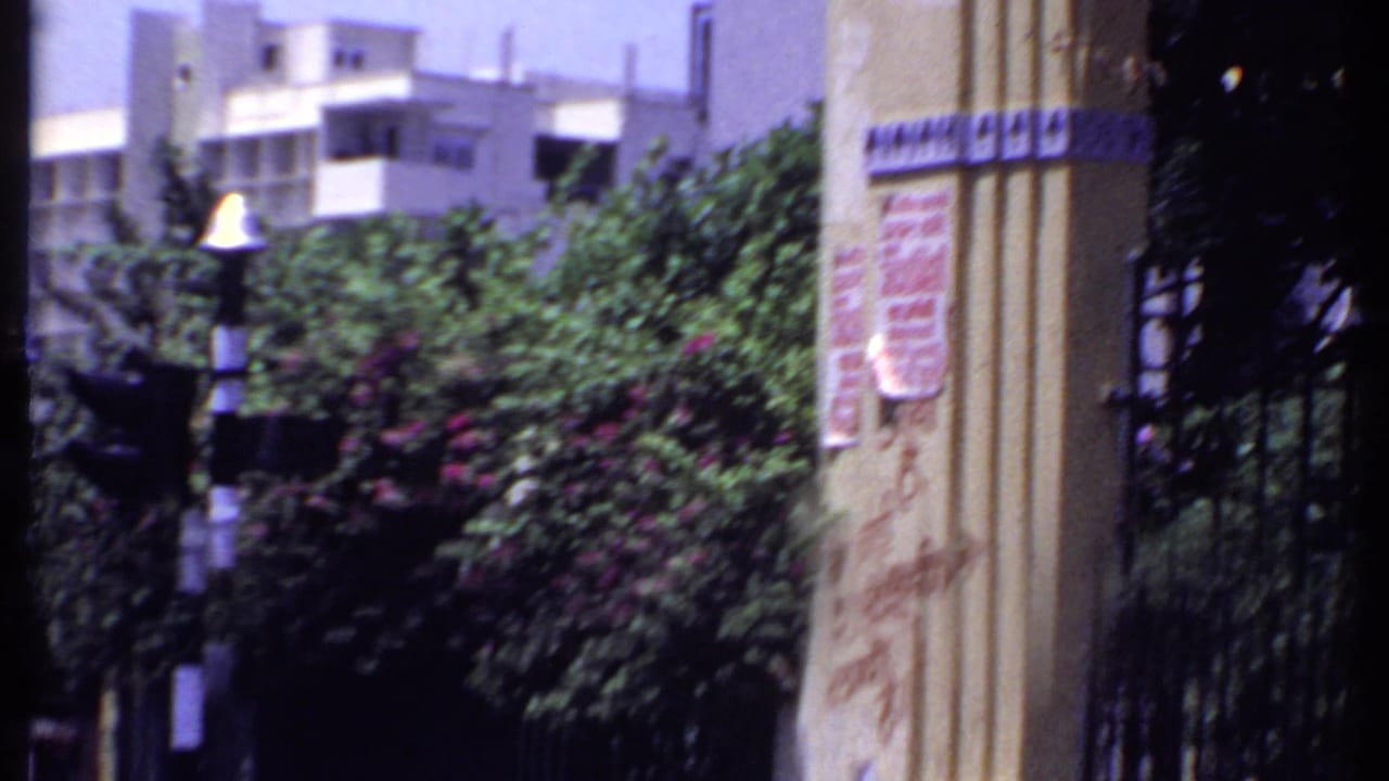 1969 India 8mm detail of a pillar with Hindi signage beside an iron gate, shrubs, striped post, and a pale building in the background.