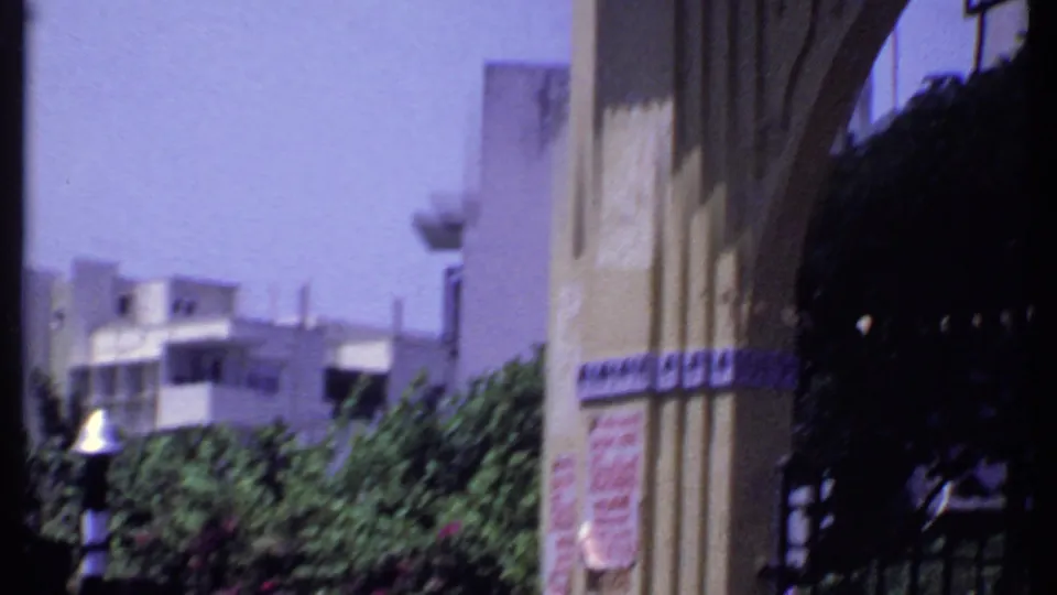 1969 India 8mm frame showing an ornate building exterior with arched detail, awning, trees, and bright blue sky.