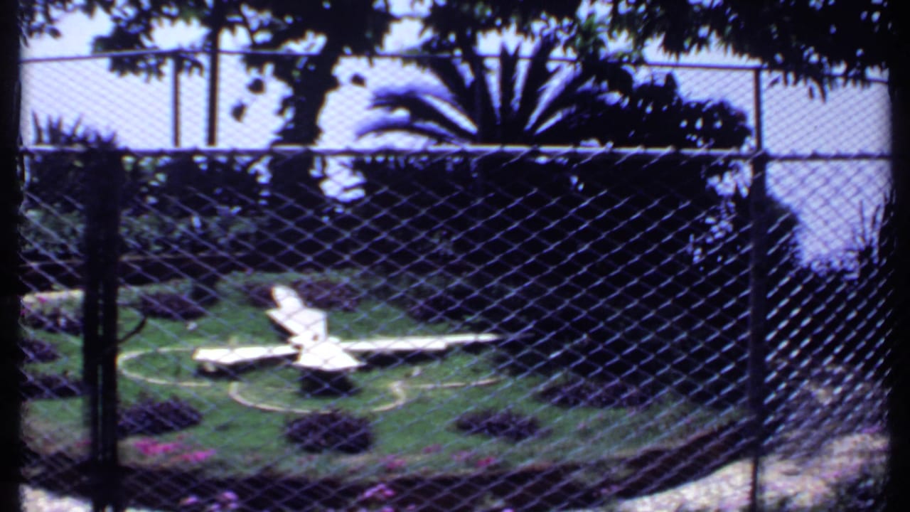 1969 India 8mm frame showing a shaded fenced lawn with white chairs, dark shrubs, pink flowers, and a curved garden edge.