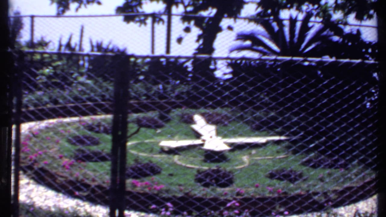 1969 India 8mm frame looking through a chain-link fence toward a landscaped lawn with white chairs, flower beds, and shaded trees.