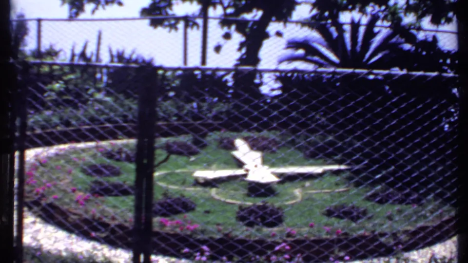 1969 India 8mm frame looking through a chain-link fence toward a landscaped lawn with white chairs, flower beds, and shaded trees.