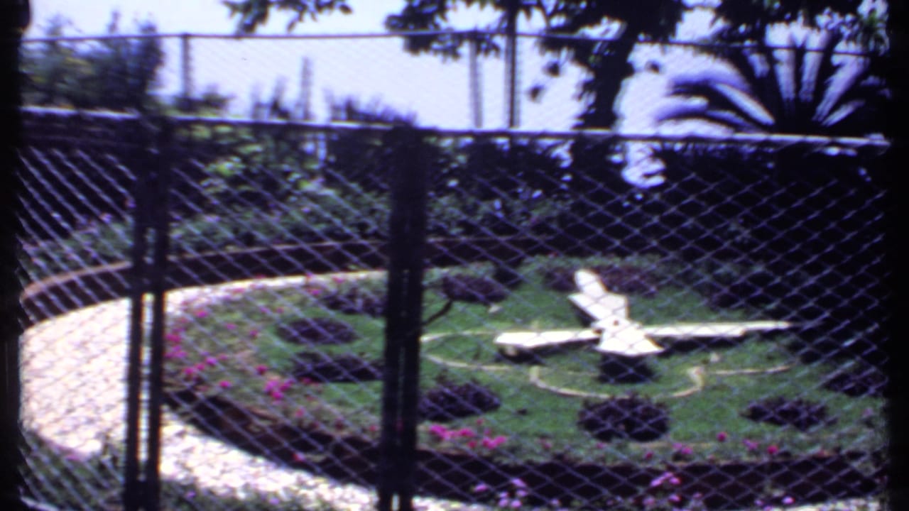 Chain-link fence before a circular garden with a white airplane sculpture and curved path.
