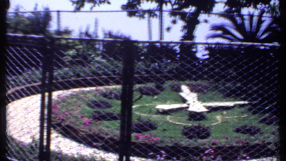 1969 India 8mm frame showing a fenced circular garden, central white airplane sculpture, lawn, flowerbeds and curved pale pathway.