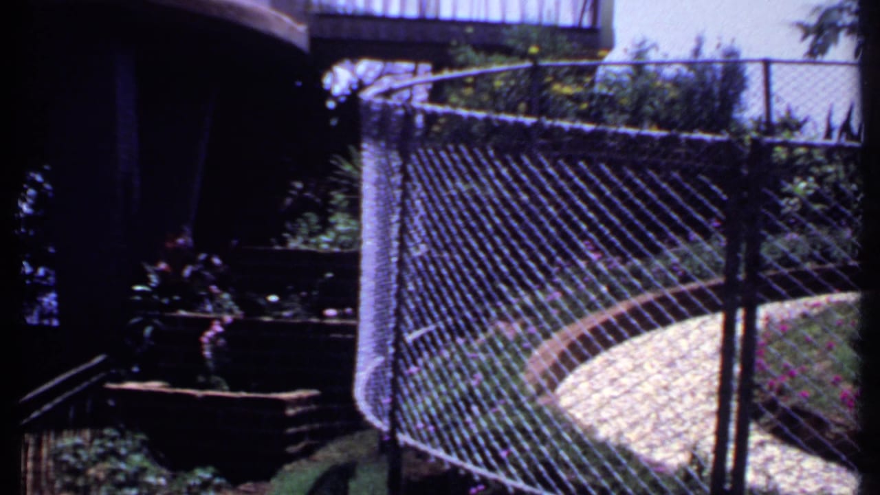 1969 India home-movie frame with the fenced garden edge, curved pale walkway, shaded foreground and planted border.