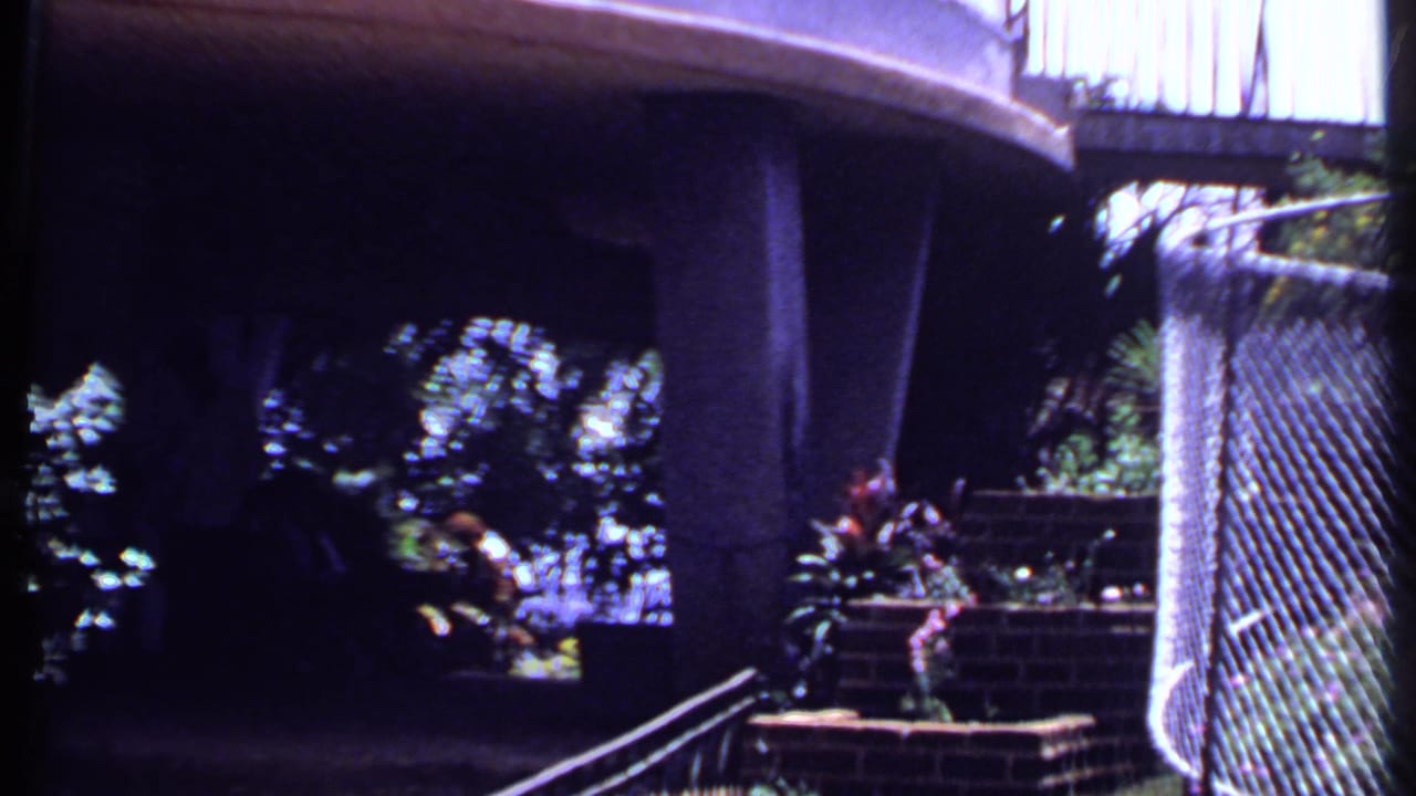 A shaded overhang or balcony foreground frames the fenced 1969 India garden, with plants and bright exterior beyond.