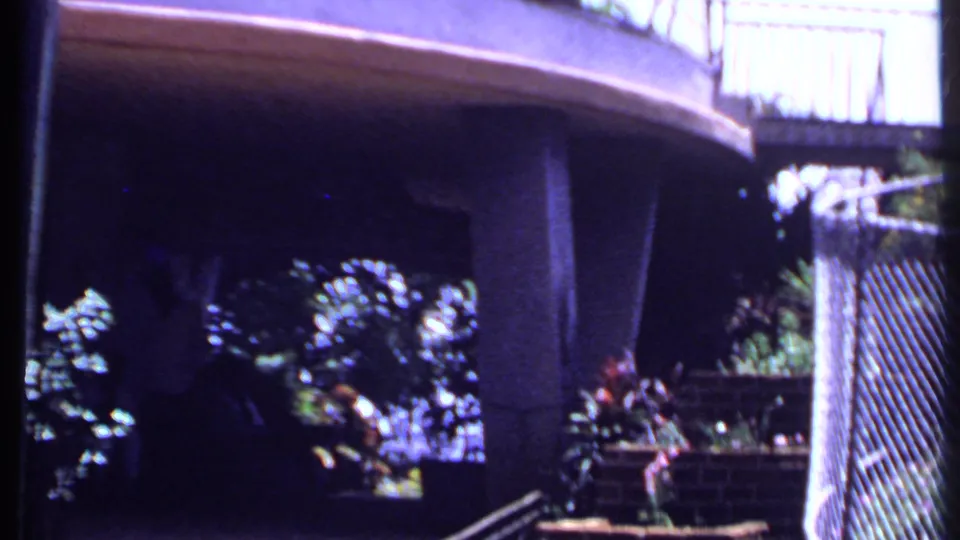 1969 India home-movie frame showing a concrete balcony or walkway structure beside tropical plants and railings.