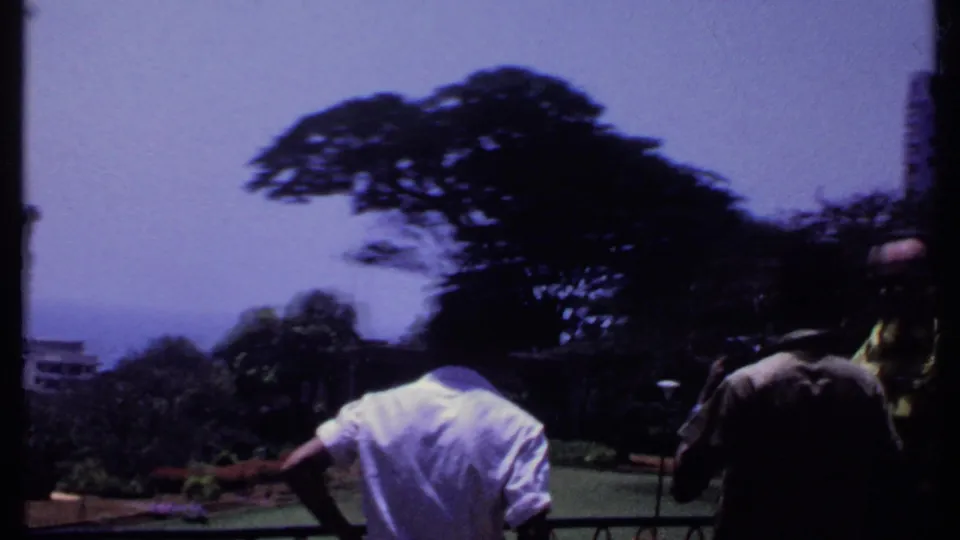 1969 India home-movie frame showing adult men on a balcony above a landscaped urban park with trees and distant high-rise buildings.
