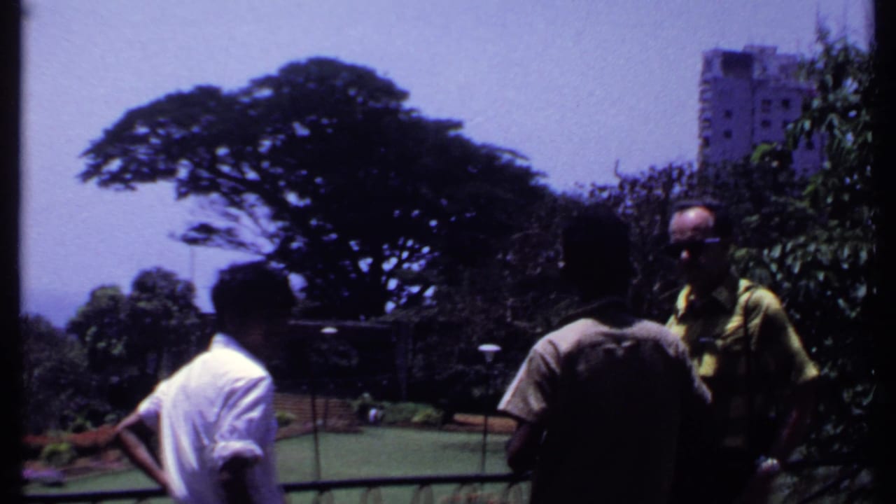 Men stand near a railing with a tree-filled park, lawn, and tall building in the background.