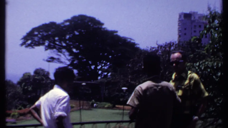 Adult visitors on a balcony or terrace look toward a green urban park in India, with a tall building visible beyond the trees.