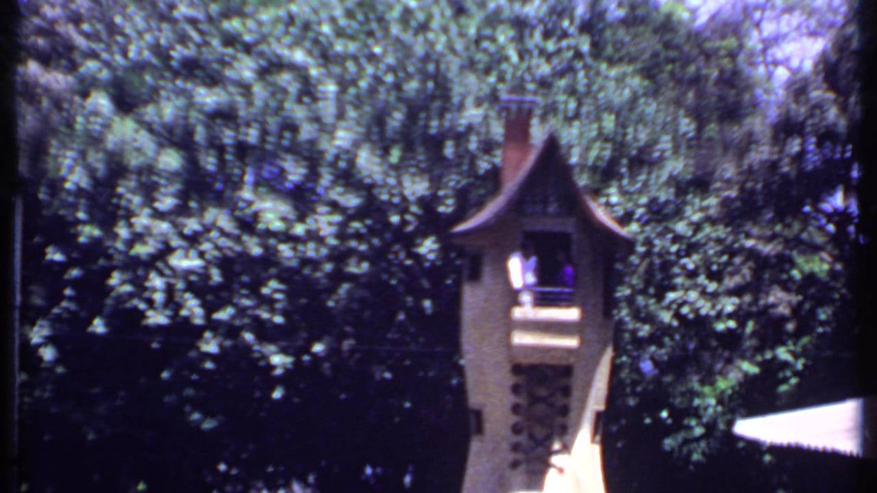 Castle-like lookout tower among dense trees with two people on a small balcony.