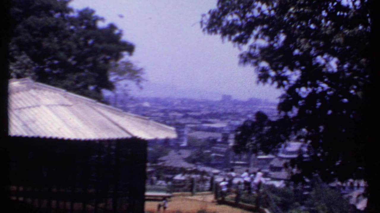 1969 India 8mm view from a lookout, showing a roof edge, visitors near railings, trees, rooftops, and a hazy town beyond.