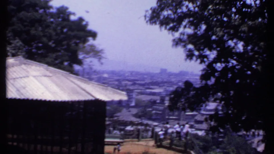 1969 India 8mm view from a lookout, showing a roof edge, visitors near railings, trees, rooftops, and a hazy town beyond.