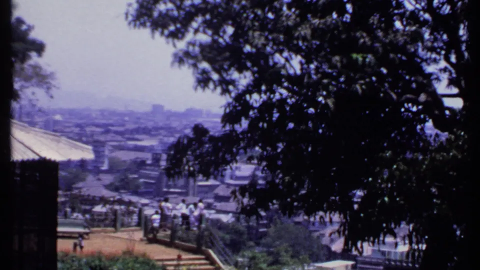 1969 India 8mm frame showing leafy trees, a terrace with distant visitors and a hazy city skyline beyond.