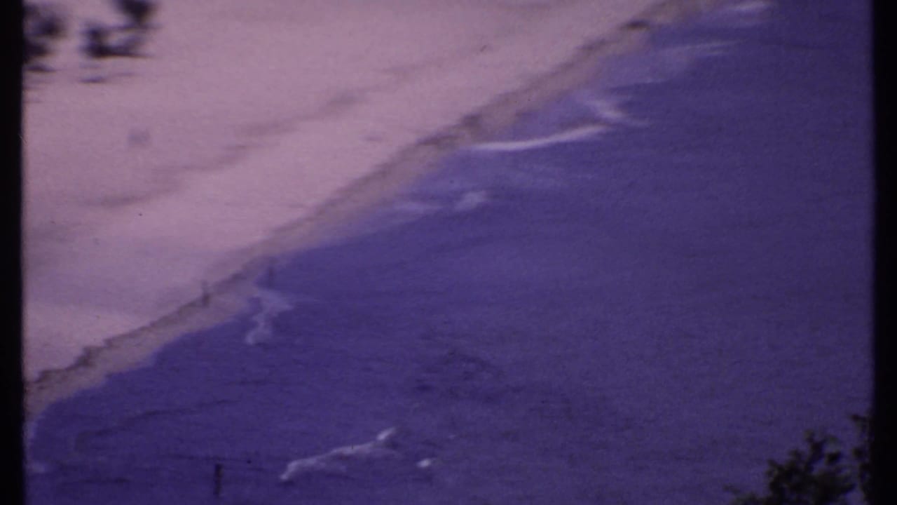 Elevated view of waves meeting a sandy beach with small figures near the shoreline.