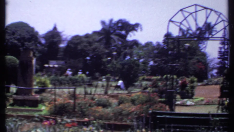 1969 India 8mm frame of a botanical garden with visitors walking among beds, palms, and a large metal arch structure.