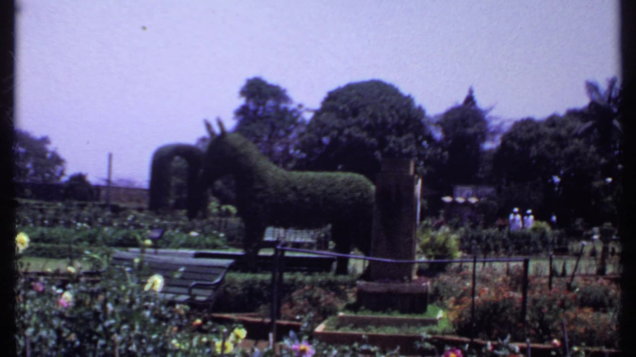Donkey-shaped topiary beside flower beds, benches, trees, and distant garden visitors.