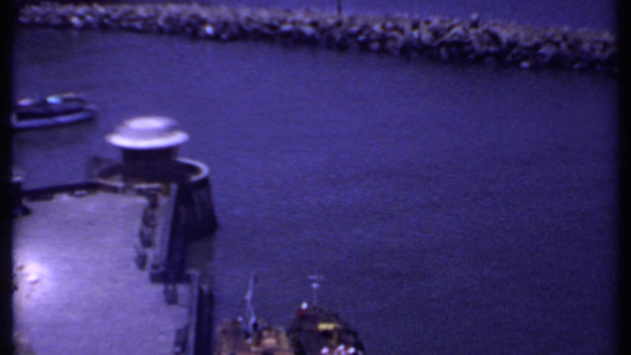 1969 India home-movie frame looking over harbor water toward a stone breakwater and small dockside boat.