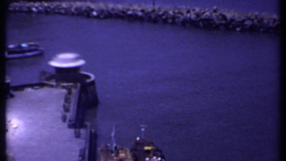 1969 India home-movie frame looking over harbor water toward a stone breakwater and small dockside boat.