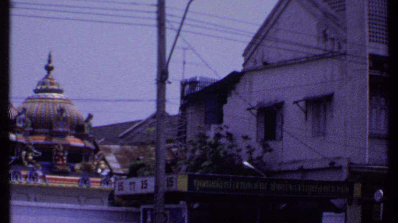 1969 8mm India street frame with a temple dome behind a white colonial-style building and Thai-script storefront signage.