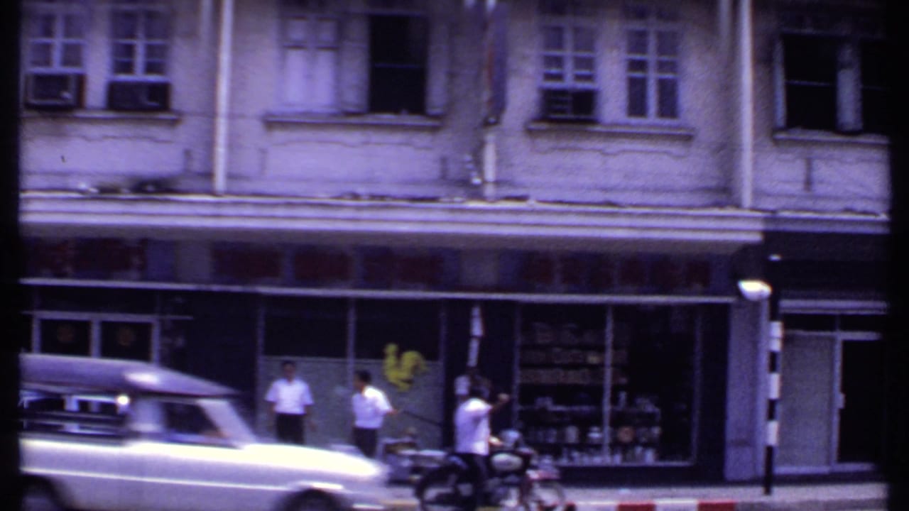 1969 8mm India street frame showing a light-colored vintage car passing pedestrians in front of an older shopfront building.