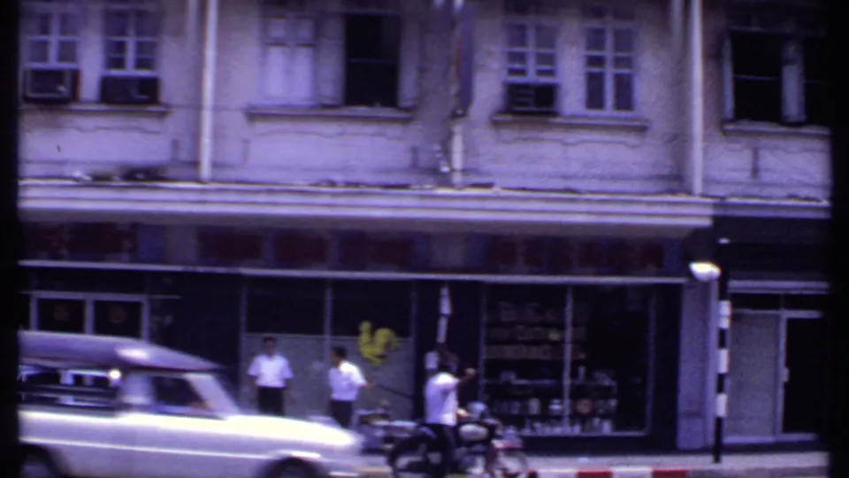 1969 8mm India street frame showing a light-colored vintage car passing pedestrians in front of an older shopfront building.